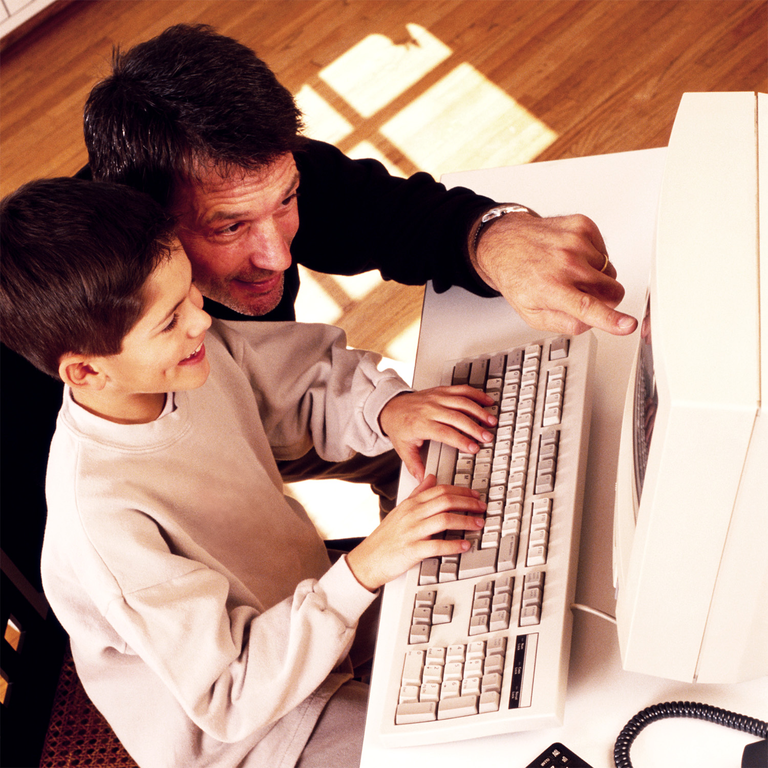 A father and young son sitting together at a desktop computer in the late 1990s, the boy typing while his dad points at the screen